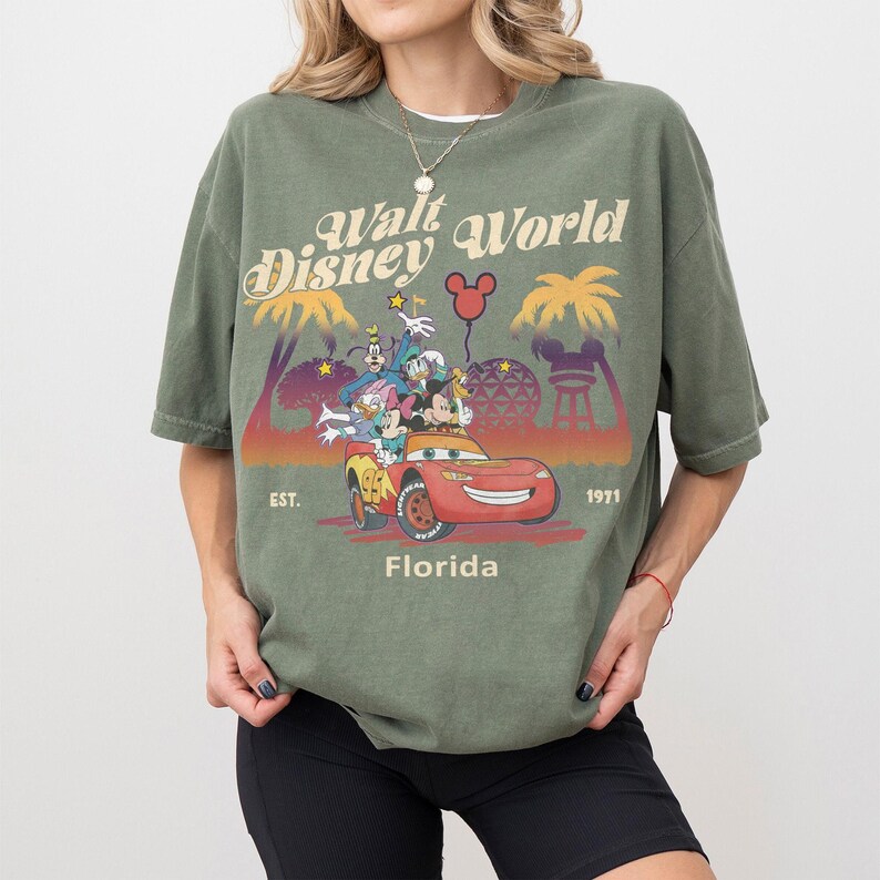 May include: An olive green t-shirt with a vintage Walt Disney World graphic. The design features cartoon characters in a red car, palm trees, and the text "Walt Disney World Florida EST. 1971".