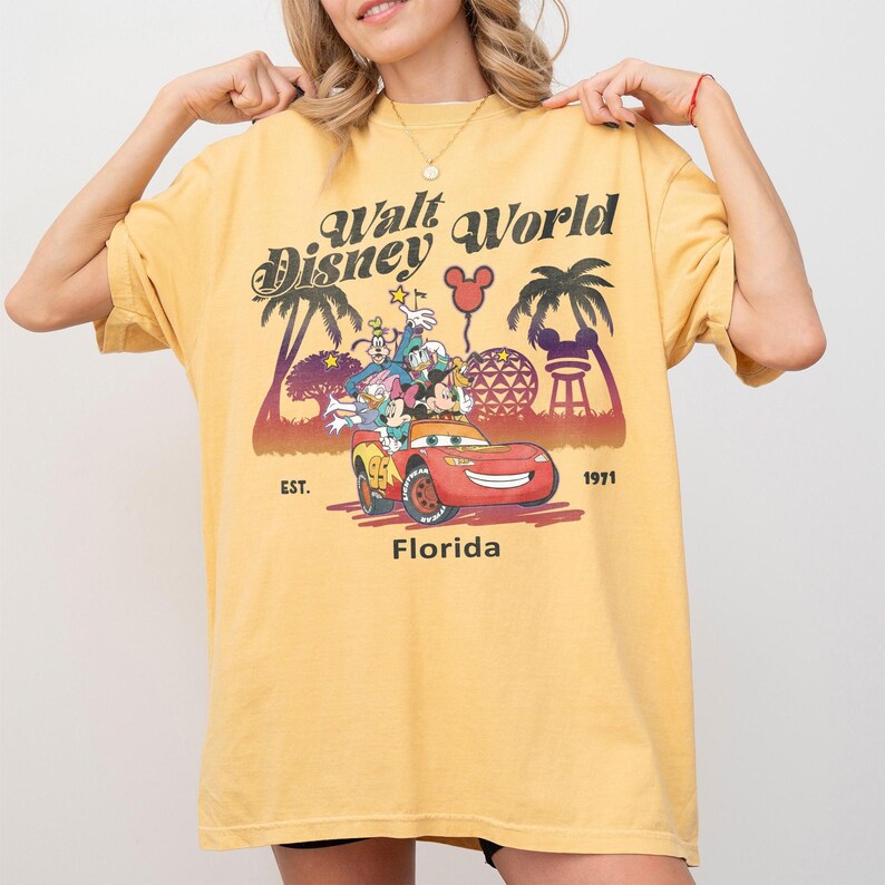 May include: Mustard yellow t-shirt featuring a retro Walt Disney World design. The graphic includes Mickey Mouse, Donald Duck, and friends in a red car, with palm trees and the text "Florida" and "1971".
