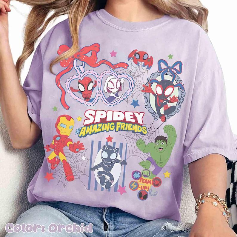 Może przedstawiać: Orchidowa koszulka z napisem "Spidey and His Amazing Friends" oraz kresk&oacute;wkowymi ilustracjami Spider-Mana, Iron Mana, Hulka i innych postaci. Projekt zawiera pajęczyny, gwiazdy i serca.