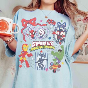 Może przedstawiać: Jasnoniebieska koszulka z napisem "Spidey and His Amazing Friends" oraz kresk&oacute;wkowymi ilustracjami Spider-Mana, Iron Mana, Hulka i innych postaci. Projekt zawiera serca, gwiazdy i pajęczyny.