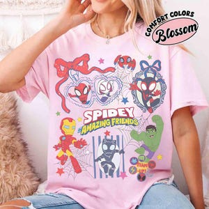 Może przedstawiać: R&oacute;żowa koszulka z napisem "Spidey and His Amazing Friends" oraz kresk&oacute;wkowymi ilustracjami Spider-Mana, Iron Mana, Hulka i innych postaci. Koszulka ma r&oacute;wnież napis "Comfort Colors Blossom".