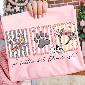 May include: Pink t-shirt featuring three illustrated panels. The panels include a Dalmatian dog, paw prints, and mouse ear headbands. The text "A little bit Devil-ish!" is written in cursive below the panels.