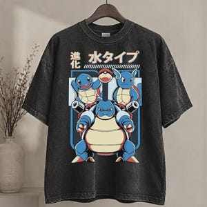 May include: A charcoal grey acid-wash t-shirt featuring a graphic print of three blue water-type creatures in various stages of evolution. The design includes Japanese text, a red and white ball icon, and a blue rectangular frame on the front.