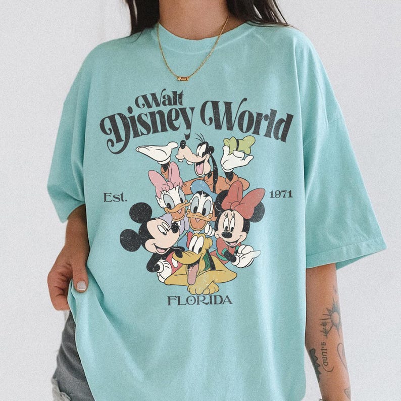 May include: A light blue t-shirt featuring the text "Walt Disney World" with the Disney characters Mickey Mouse, Minnie Mouse, Donald Duck, Daisy Duck, Goofy, and Pluto. The shirt also includes the text "Est. 1971" and "Florida".