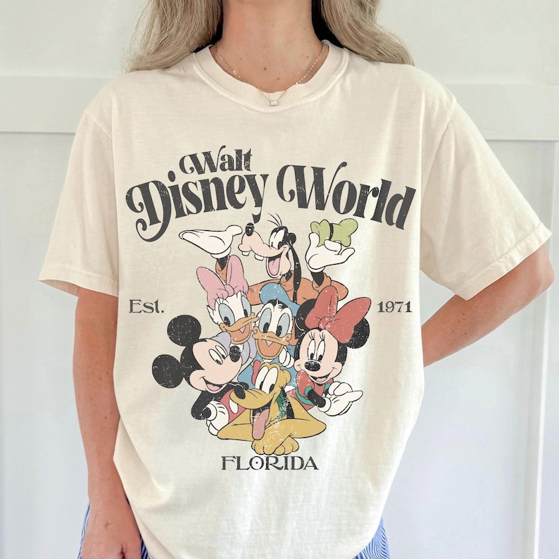 May include: Cream-colored t-shirt featuring a graphic of Disney characters, including Mickey Mouse, Minnie Mouse, Donald Duck, and Goofy. The shirt has the text "Walt Disney World" and "Est. 1971 Florida" in a vintage style.
