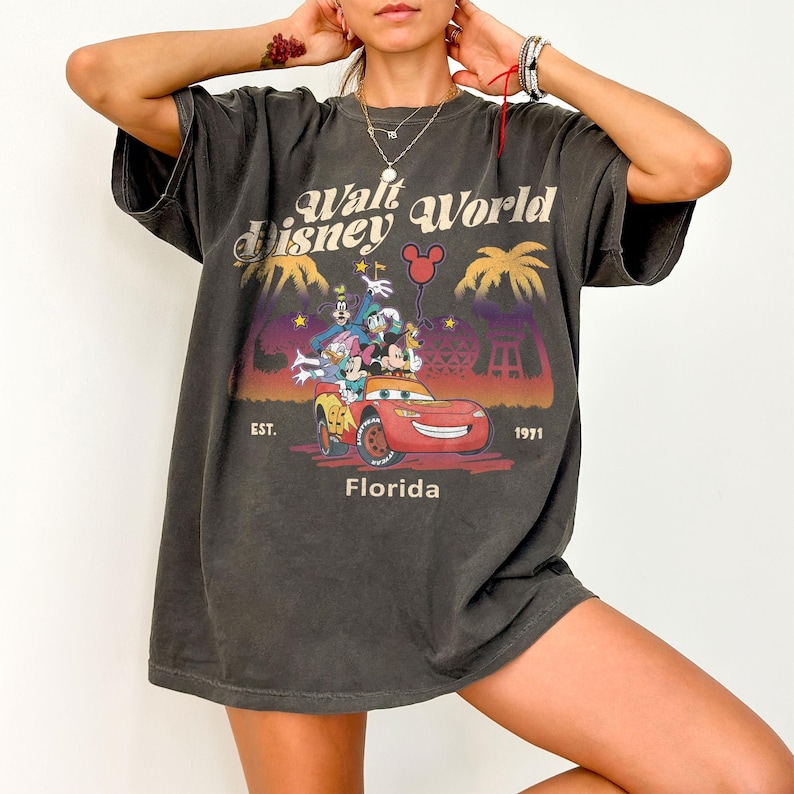 May include: Dark grey t-shirt with a colourful graphic of Disney characters in a red car, the text reads "Walt Disney World Florida" and "EST. 1971". The shirt has a vintage, worn appearance.