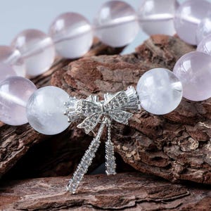 May include: A beaded bracelet featuring large, round, pale pink and clear quartz beads. The bracelet is accented with a silver-colored bow-shaped charm, embellished with small, sparkling stones, and dangling accents. The bracelet is displayed on a textured, brown surface.