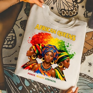 May include: A cream-colored sweatshirt featuring the text "AFRICAN QUEENS" in yellow. The graphic depicts three women wearing colorful head wraps and jewelry, with the words "POWER. BEAUTY. STRENGTH." below.