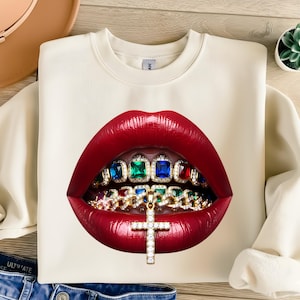 Puede incluir: Sudadera de color crema con un gráfico de labios rojos, una parrilla enjoyada y un colgante de cruz dorada. La parrilla tiene gemas verdes, azules y rojas. La sudadera tiene mangas largas y cuello redondo.
