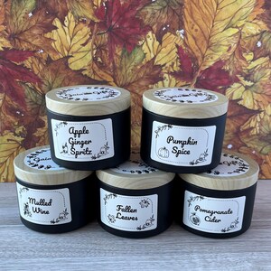May include: Five black scented candles with wooden lids are displayed. Each candle has a white label with a different scent name: Apple Ginger Spritz, Pumpkin Spice, Mulled Wine, Fallen Leaves, and Pomegranate Cider. The background features autumn leaves.