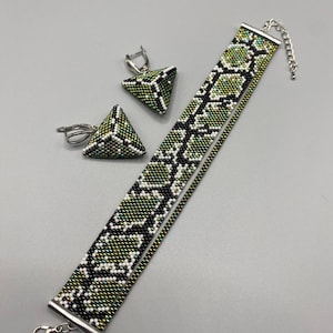 Könnte beinhalten: Ein perlenbesetztes Choker-Halsband und ein Paar passende dreieckige Ohrringe mit einem grün-schwarz-weißen Schlangenhautmuster. Das Schmuckset enthält silberfarbene Metallbeschläge, einen Karabinerverschluss und eine verstellbare Kette.