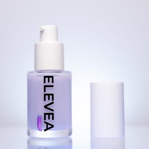 May include: A lavender-colored pump bottle with a white cap and the word "ELEVEN" printed vertically in black. A purple brushstroke design is below the text. The bottle's cap is separate, and the background is a gradient of white and light blue.