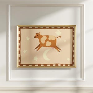 May include: A framed art print featuring a whimsical illustration of a brown and white cow leaping across a light peach background. The artwork includes a crescent moon, stars, and a sun-like shape. The frame is gold.