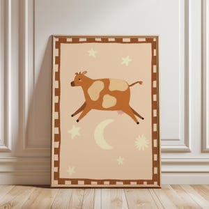 May include: A framed art print featuring a whimsical illustration of a brown and white cow leaping across a light peach background. The artwork includes stars, a crescent moon, and a decorative border, creating a playful, dreamy aesthetic.