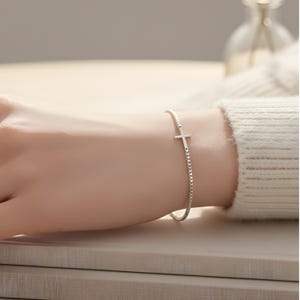 May include: A delicate silver bracelet featuring a small cross charm. The bracelet has a thin, textured chain and is worn on a wrist. The cross is centered on the wrist, and the bracelet is simple and elegant.