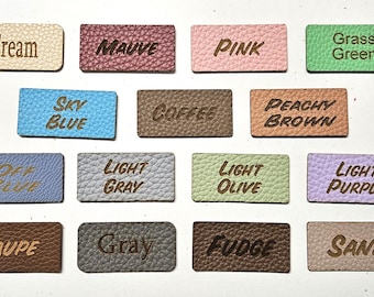 Könnte beinhalten: Eine Sammlung von fünfzehn rechteckigen Kunstlederetiketten mit genarbter Textur. Jedes Etikett hat eine einzigartige Farbe wie Creme, Mauve, Pink, Grasgrün, Himmelblau, Kaffee, Pfirsichbraun, Off-Blue, Hellgrau, Helloliv, Helllila, Taupe, Grau, Fudge und Sand, wobei der entsprechende Farbname in dunkelbrauner Schrift eingraviert ist.