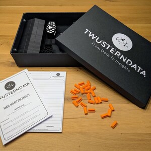 Può includere: Un set di scatole nere con il testo "TWÜSTERNDATA From Data To Insights" e un logo. All'interno della scatola ci sono componenti neri e un oggetto bianco e nero. Ci sono pezzi arancioni sparsi sulla superficie di legno, insieme a un libretto e un blocco note.