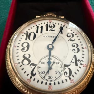 May include: A gold-toned Hamilton pocket watch with a white face, black numerals, and a smaller seconds dial. The watch is in a red velvet case, showcasing its intricate design and classic appeal.