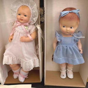 May include: Two vintage Effanbee baby dolls in their original boxes. One doll wears a pink dress and bonnet with a pink ribbon. The other doll is in a blue gingham dress and a blue headband. Both dolls have painted features.