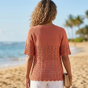 Könnte beinhalten: Ein terrakottafarbenes Häkeloberteil mit einem fest gestrickten oberen Bereich und einem dekorativen Lochmuster an der unteren Hälfte und den Ärmeln. Das Kleidungsstück hat kurze Ärmel mit gewellten Kanten sowie einen runden Halsausschnitt.