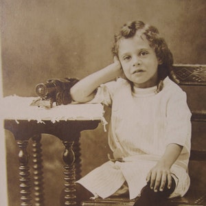 May include: A sepia-toned photograph of a young child sitting on a chair with a toy train on a table beside them. The child is wearing a white dress and has their hand on their chin. The photo is labelled "Proof".