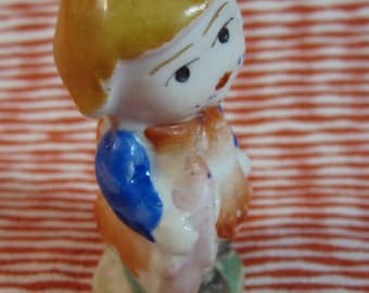 Puede incluir: Una pequeña figurita de porcelana de un niño con cabello rubio, que lleva un vestido azul y naranja. La figurita está de pie sobre una base con un diseño verde y marrón.
