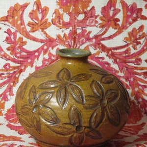 Könnte beinhalten: Eine runde, braune Keramikvase mit geprägten Blumenmustern. Die Vase hat einen schmalen Hals und eine leicht strukturierte Oberfläche. Der Hintergrund zeigt einen gemusterten Stoff mit orangefarbenen und rosa Blumenmotiven.