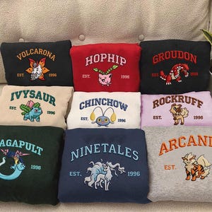 May include: A collection of embroidered sweatshirts in various colors, each featuring a different Pokémon character and the year 1996. The sweatshirts have the Pokémon's name embroidered above the character.