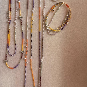Könnte beinhalten: Eine Sammlung von Halsketten und Armbändern mit Perlen in verschiedenen Farben. Die Halsketten haben orange, gelbe, lila und silberne Perlen, einige mit kleinen perlenartigen Perlen. Die Armbänder haben ähnliche Farbschemata und Designs.