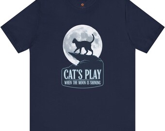 Pode incluir: Camiseta azul marinho com um gráfico de um gato preto contra uma lua cheia. O texto "CAT'S PLAY WHEN THE MOON IS SHINING" está abaixo. Uma camiseta casual de manga curta.
