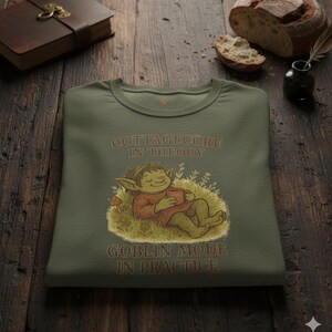 Puede incluir: Sudadera verde oliva con cuello redondo y el texto "COTTAGECORE IN THEORY, GOBLIN MODE IN PRACTICE". El gráfico muestra un duende de dibujos animados con una taza. La sudadera está doblada sobre una superficie de madera.