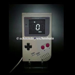 Könnte beinhalten: Eine digitale Uhr im Retro-Stil, die einem klassischen Handheld-Spielgerät nachempfunden ist. Die Uhr hat eine grau-weiße Farbgebung mit einem schwarzen Bildschirm, der die Zeit in weißen Ziffern anzeigt. Sie hat ein kreuzförmiges Steuerkreuz, rosa Knöpfe und die Schriftzüge "SELECT" und "START".