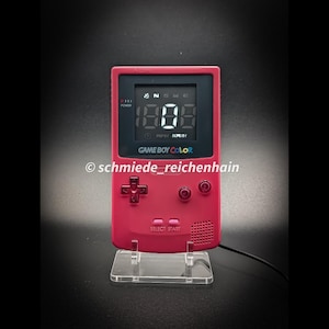 Könnte beinhalten: Eine beerenfarbene Game Boy Color Handheld-Konsole, die zu einer Digitaluhr umgebaut wurde. Der Bildschirm zeigt die Uhrzeit 18:08 in weißen Digitalziffern. Das Gerät verfügt über ein Steuerkreuz, zwei Tasten und den Text schmiede_reichenhain.