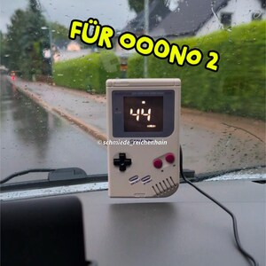 Può includere: Una console di videogiochi portatile color crema con un pad direzionale nero, pulsanti rosa e un display digitale che mostra il numero 44. Il testo "FÜR OOOONO 2" è in giallo sopra la console.