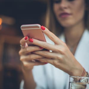 Pode incluir: Um smartphone rosa dourado é segurado com as duas mãos com esmalte vermelho. A pessoa está usando uma blusa branca e uma pulseira. Um copo de água está em primeiro plano.