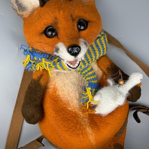 Puede incluir: Bolso de fieltro con forma de zorro naranja, con una bufanda a rayas azules y amarillas y un ganso blanco. El bolso tiene una correa marrón y está diseñado para llevarlo al hombro. El zorro tiene ojos grandes y expresivos.