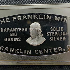 May include: A rectangular silver ingot from The Franklin Mint featuring a portrait of Benjamin Franklin. The text reads: THE FRANKLIN MINT, GUARANTEED 500 GRAINS, SOLID STERLING SILVER, and FRANKLIN CENTER, PA. The central design is set against a black background.