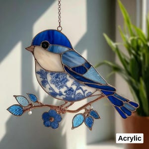 May include: A stained glass bird suncatcher in shades of blue and white, perched on a bronze-colored branch with blue leaves and flowers. The bird's body features a floral pattern. The word "Acrylic" is in the bottom right corner.
