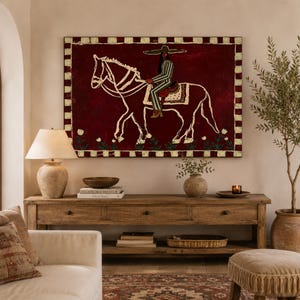 May include: A rectangular wall art piece featuring a dark red background with a white outline illustration of a person in a wide-brimmed hat riding a horse. The border consists of alternating cream and dark red squares with small floral motifs.