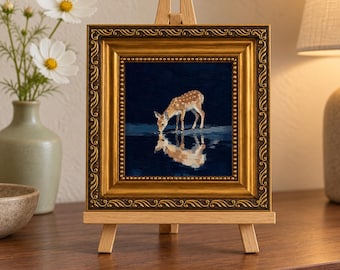 May include: A small square painting of a spotted fawn drinking from a dark pool of water, creating a clear reflection. The artwork is encased in an ornate, metallic gold frame and is displayed on a light-colored wooden tabletop easel.