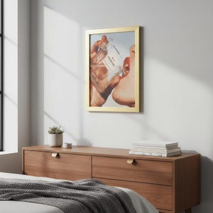 May include: A framed print featuring a person drinking from a Chanel No. 5 perfume bottle. The image is in a gold frame and is displayed above a wooden dresser. The person's lips are painted red.