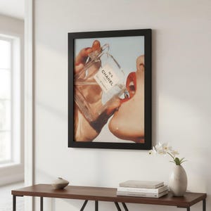 May include: Framed print featuring a close-up of a person's face with red lipstick, drinking from a Chanel No. 5 perfume bottle. The print is in a black frame and hangs on a white wall. A wooden table with a vase and books is below the print.