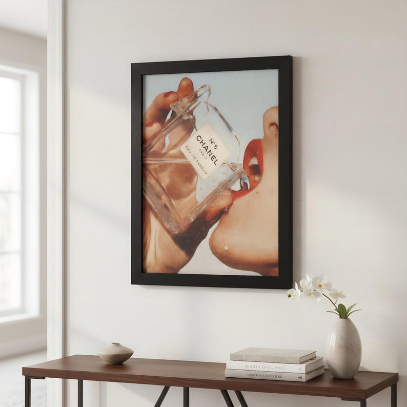 May include: Framed print featuring a close-up of a person's face with red lipstick, drinking from a Chanel No. 5 perfume bottle. The print is in a black frame and hangs on a white wall. A wooden table with a vase and books is below the print.
