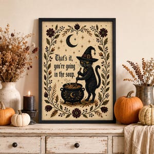Può includere: Una stampa artistica incorniciata raffigurante un gatto nero con un cappello da strega che mescola un calderone con il testo That's it, you're going in the soup. L'illustrazione è circondata da un bordo floreale decorativo con luna crescente e stelle.