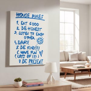 May include: A white canvas with blue handwritten text that reads "HOUSE RULES" followed by seven rules for a household. A small white lamp, a succulent in a white pot, and a stack of books sit on a wooden table.