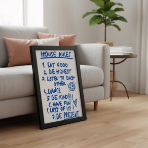May include: A framed sign with the words "HOUSE RULES" written in blue marker. The rules include "Eat Good," "Be Honest," "Listen to Each Other," "Dance," "Be Kind," "Have Fun," and "Be Present." The sign is propped against a gray couch.