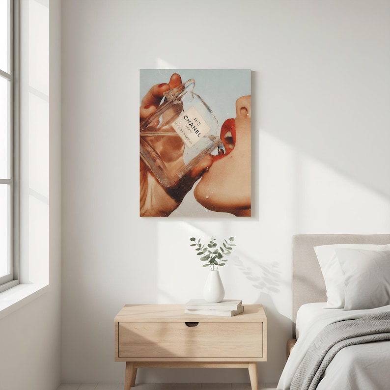 May include: A canvas print depicts a person's face with red lips drinking from a Chanel No. 5 perfume bottle. The bottle is clear with a white label. The artwork is displayed on a white wall above a wooden nightstand.