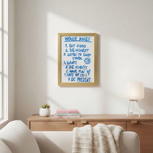 May include: A framed print with the words "HOUSE RULES" in blue ink. The rules include "EAT GOOD," "BE HONEST," and "BE PRESENT." The frame is gold and hangs on a white wall. A small white lamp and a succulent in a white pot are visible.
