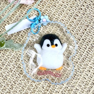 Puede incluir: Un llavero con forma de pingüino con un borde transparente y festoneado. El pingüino tiene la cabeza negra, el cuerpo blanco y las patas naranjas. El texto "Oh Squish Me" está impreso en el plástico transparente. Una flor azul y un llavero están adjuntos.