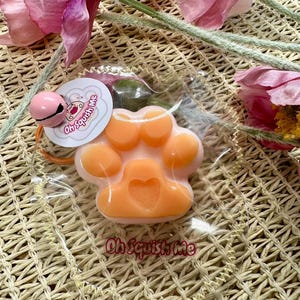 May include: A peach-coloured paw-shaped soap with a white base is encased in clear plastic. A pink bell and a tag with the text "Oh Squish Me" are attached. The soap has a heart-shaped centre. The background includes pink flowers.